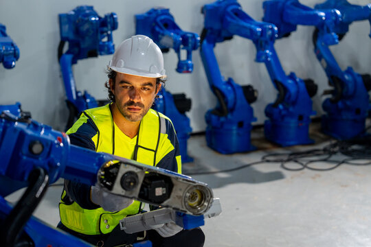 Engineer in yellow vest and white helmet inspects blue robotic arm in industrial workshop with focused intensity