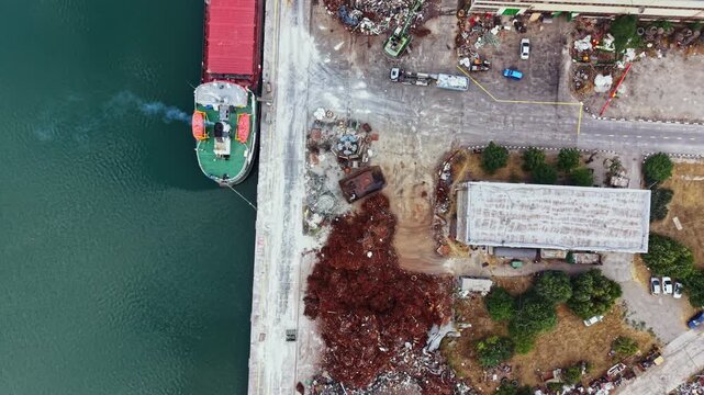 A cargo ship is docked along a waterfront. Nearby, there are piles of scrap materials and a storage building along the shore. The area shows industrial activity and boats.