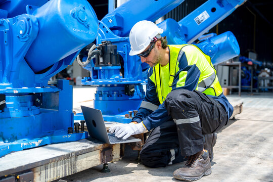 Engineer in safety gear kneels beside blue industrial machinery using laptop under bright workshop lighting
