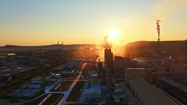 Smoke rises from multiple factories during sunset in an industrial area. The sun sets behind the hills, casting light over buildings. Machinery is visible in the foreground.