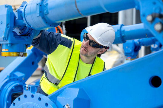 Engineer in safety gear adjusts blue robotic arm under bright industrial lighting