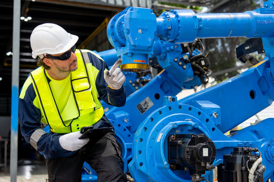 Engineer in high-visibility gear points at blue robotic arm joint while holding tablet in workshop