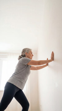 Senior woman performing standing wall push exercise inside bright home, authentic active aging concept showing independence confidence value value value value value value value value