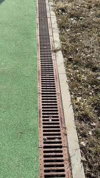 Closeup sports field drainage grate showing rusted iron slots, debris lodged along turf edge, concrete border, textured artificial