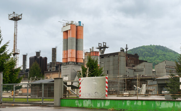Industrial complex  with orange and gray storage silos, metal production halls, chimneys, pipelines and security fencing. Forested hills and cloudy weather contrast with the aging heavy-industry infra