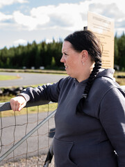 Side profile portrait of a smiling woman leaning on a fence at an outdoor go-kart track on a sunny summer day. Natural lifestyle image with blurred forest background and copy space. © HelenaUpNorth
