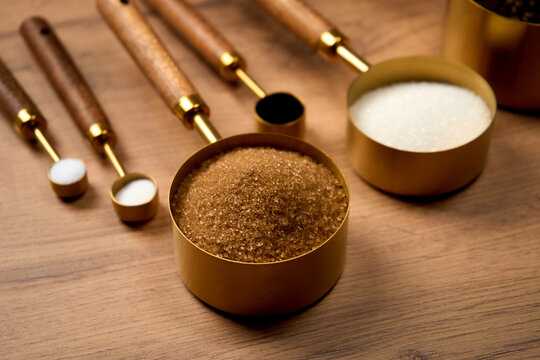 Baking ingredients brown sugar in a measuring spoon; other measuring spoons containing salt, sugar, and chocolate lie nearby on a wooden kitchen table. Chicken eggs on a stand are also close by.