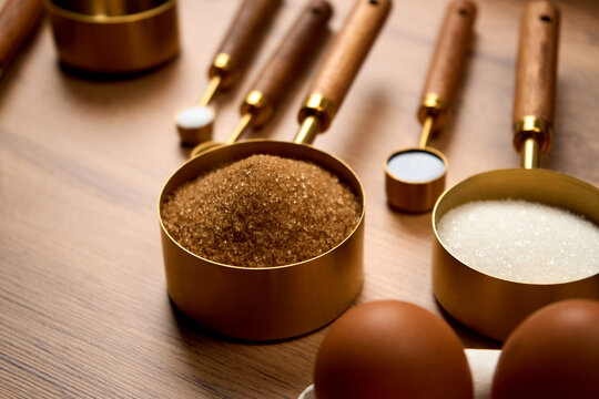 Baking ingredients brown sugar in a measuring spoon; other measuring spoons containing salt, sugar, and chocolate lie nearby on a wooden kitchen table. Chicken eggs on a stand are also close by.