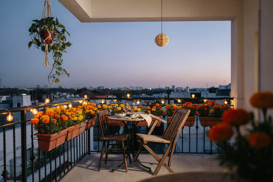 A cozy balcony decorated with glowing string lights potted orange marigold flowers a small table and two wooden chairs overlooking a serene cityscape at twilight offering a peaceful evening retreat