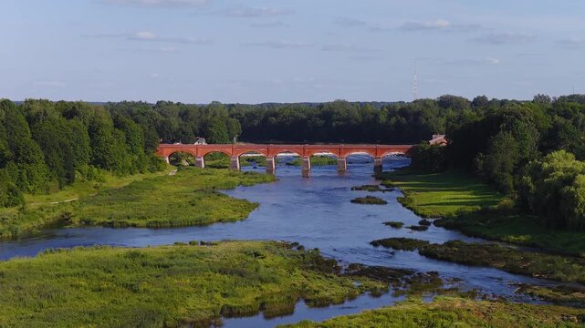Aerial drone shot shows Venta River under the red brick arched bridge in Kuldiga, Latvia, gliding downstream to the Venta waterfall, with summer light and wide view.