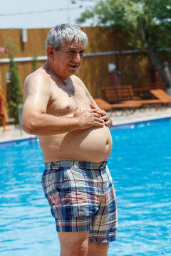 Man stands by pool, bare-chested, smiling. Wears plaid swim trunks, hands on belly.