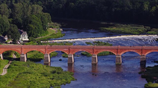 Aerial view shows the red brick arched bridge over the Venta River in Kuldiga, Latvia, with Ventas Rumba flowing behind, late day light, people strolling.