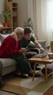 Caucasian young adult man teaching senior mother or grandmother how to use laptop computer while sitting on sofa. Ideal for digital literacy campaigns or senior care services, vertical