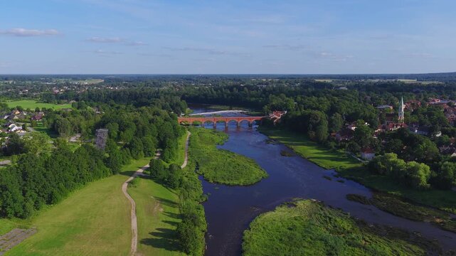 Aerial view over Kuldiga, Latvia, tracks the Venta River to the red brick bridge and Venta Rapid. The camera glides downstream past parkland, paths, houses, and a church.