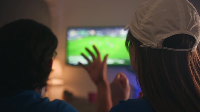 Couple sitting on a sofa at home watching a football match on TV. During a goal celebration they raise beer bottles and toast while cheering for their favorite team. Sports fandom, victory and shared