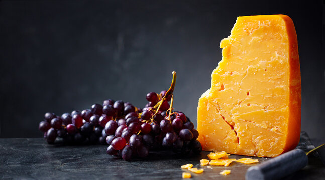 Piece of aged cheddar cheese with red grapes on dark background with copy space. Close up.