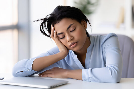 African American woman resting her head on her hand while sitting at a desk with a laptop and plant in a bright office environment
