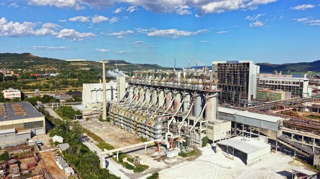 A large factory is seen with multiple silos and machinery. The scene shows activity around the buildings with smoke rising. It is daytime with a clear sky and some clouds.