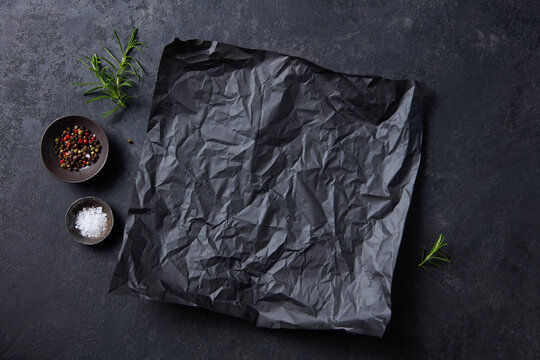 Black parchment paper, spice bowls and rosemary sprigs flat lay. Dark grey background. Copy space, close up. Top view.