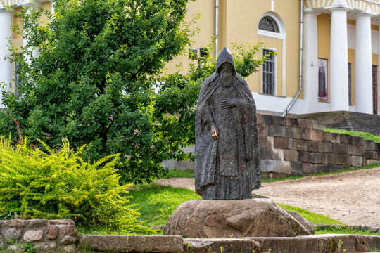 Ostashkov, Tver region, Russia - July 26, 2026: Majestic bronze statue of hooded Orthodox monk stands on large stone base in front of historic yellow church building