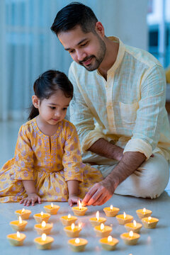 Father and daughter lighting traditional oil lamps together during Diwali celebration, surrounded by glowing candles on a bright indoor floor