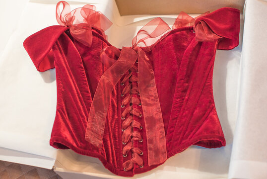 Luxurious deep red velvet corset top with off-the-shoulder straps and ribbon bows hanging on a hanger or lying in a gift box. Elegant feminine fashion lingerie garment with lacing detail