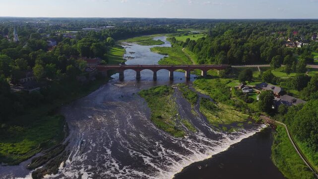 Aerial view of Kuldiga, Latvia shows Venta River, Venta Rapid, and the red brick arched bridge, with cars crossing, foamy currents, and braided channels in sun.