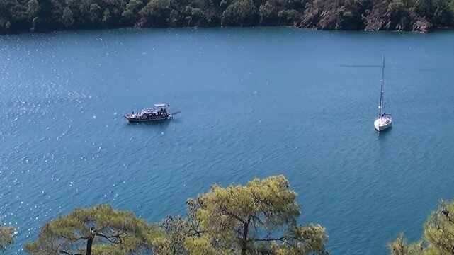 Boats glide peacefully on Oludeniz Bay's vibrant turquoise waters in Turkey. A serene sailboat and lively motorboat traverse the stunning expanse, flanked by lush, verdant trees.