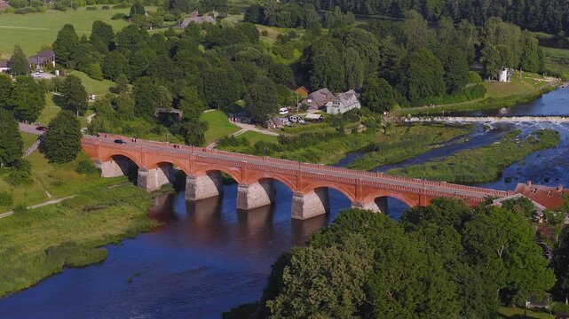 Aerial view shows Kuldiga, Latvia, red brick arched bridge and Venta Rapid as cars and pedestrians move, water flows over the weir, and a smooth drone pan glides