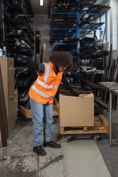 A female worker in a warehouse lifts a heavy cardboard box and shows signs of back pain. She wears safety gear while handling the load.