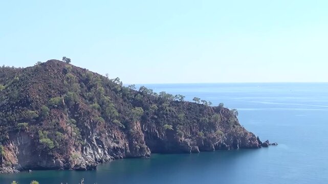 A magnificent view of the Fethiye coastline in Turkey. Rugged cliffs, adorned with lush green trees, majestically meet the calm, sparkling blue Mediterranean Sea on a bright day.