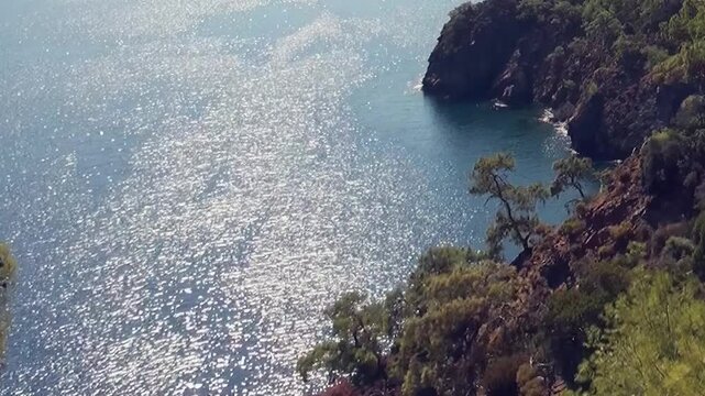 Golden sunlight dances across the serene waters near Fethiye Turkey. Lush green foliage clings to the rugged cliffs, overlooking the shimmering turquoise sea on a bright, beautiful day.