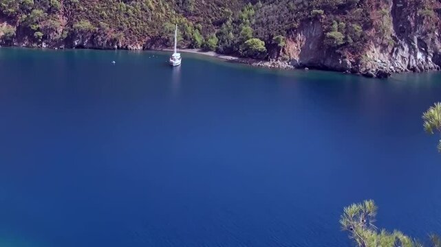 Witness the breathtaking beauty of Oludeniz Bay, Turkey, where a serene sailboat rests peacefully on vibrant azure waters. Lush green landscapes and towering cliffs frame this idyllic daytime vista.