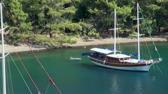 A majestic wooden gulet floats peacefully on the clear turquoise waters of Oludeniz Bay Turkey. Lush green trees line the tranquil shore, creating an idyllic summer paradise.