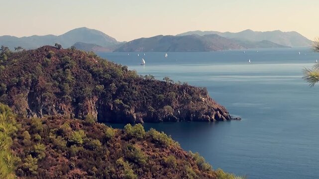 Witness the majestic beauty of Oludeniz Bay in Turkey. White sails glide peacefully over crystal waters near a verdant peninsula. Distant mountains frame this idyllic, tranquil daytime scene.