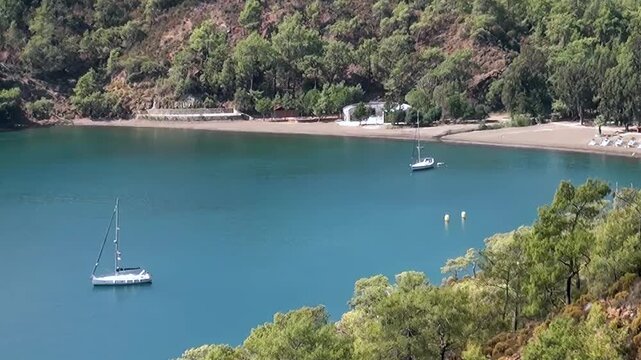 A tranquil daytime vista of Oludeniz Bay, Turkey. Majestic sailboats gracefully anchor in crystal blue waters, framed by verdant hills and a peaceful sandy shoreline. Perfect for relaxation.
