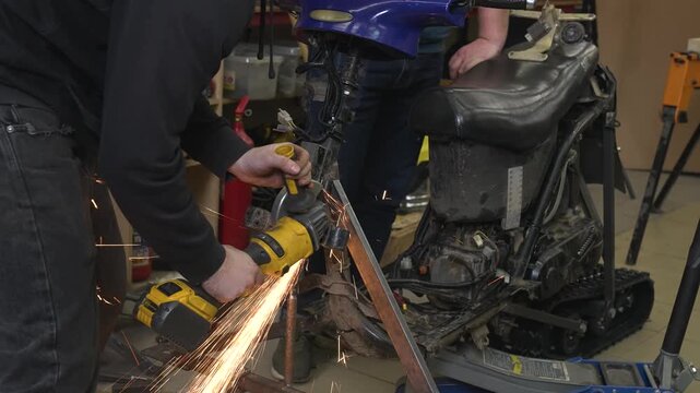 A worker uses a grinder to cut off excess parts from a snowmobile's metal frame, sparks flying in all directions.