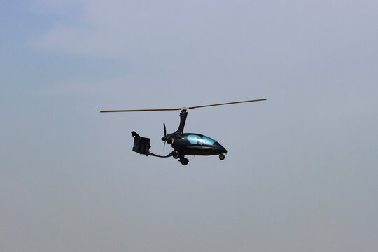 Autogyro in flight with its overhead rotor spinning against a cloudy gray sky