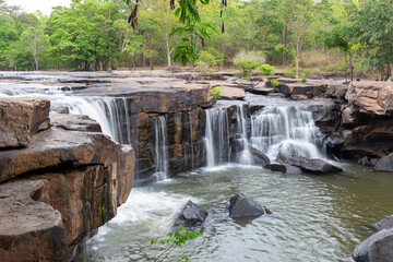 Scenic waterfall cascading over rocky stone ledges into a natural river pool surrounded by lush green forest © wannasak