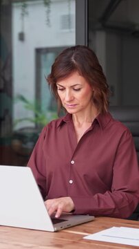 Latin mature businesswoman working on laptop in modern office. Smiling woman entrepreneur runs online business from coworking interior. Female company founder sits at workplace desk. Vertical