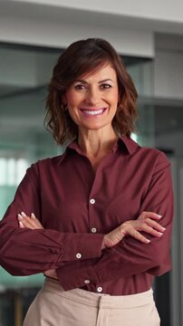 Crossing arms and smiling, hispanic mature businesswoman poses standing in office portrait. Happy woman company owner looks at camera. Successful female runs business with confidence. Vertical