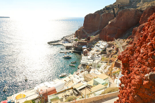 The fishing harbor of Ammoudi Bay lies beneath red volcanic cliffs in Santorini, Greece, its white buildings and boats glowing in Mediterranean sunlight.