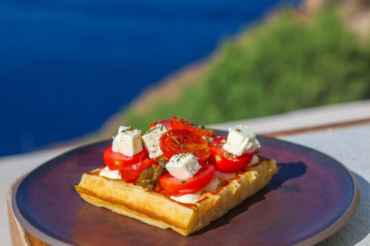 A savory waffle topped with fresh tomato, feta cheese, and capers sits on a plate overlooking the Aegean Sea in Santorini, Greece, blending local flavor and coastal charm.