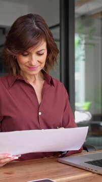 Latin businesswoman checking paper report at desk in office. Mature woman financial analyst holds document, works on laptop. Female expert reviews business contract at workplace. Vertical