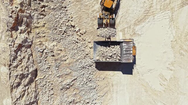 A large machine loads rocks into a truck at a quarry. The scene shows the tools and equipment used for extracting stone. The location is busy with activity as work proceeds in the daytime.