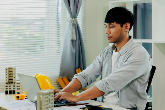 Asian engineer double-checking project specs using laptop and calculator beside architectural model.