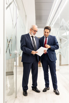 Vertical photo of two people between 40 and 60 years old, dressed in suits, talking in the middle of a hallway with papers in a modern office. Concept of lawyers or businesspeople.