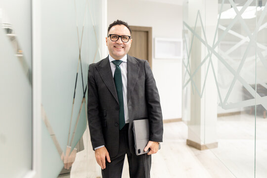 A lawyer or businessman in his fifties, wearing a suit, looks happily at the camera with a tablet in his hand in a modern office. Concept of office staff or a professional in a suit.