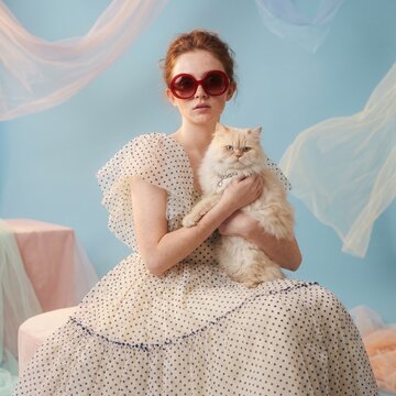 Stylish Redhead Woman in Polka Dot Dress and Red Oval Sunglasses Holding Fluffy White Cat in Studio