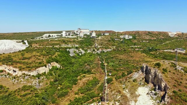 The scene shows an industrial area with buildings, machinery, and towers located on a hillside. The land is mostly green and includes some rocky sections. It is bright and sunny.